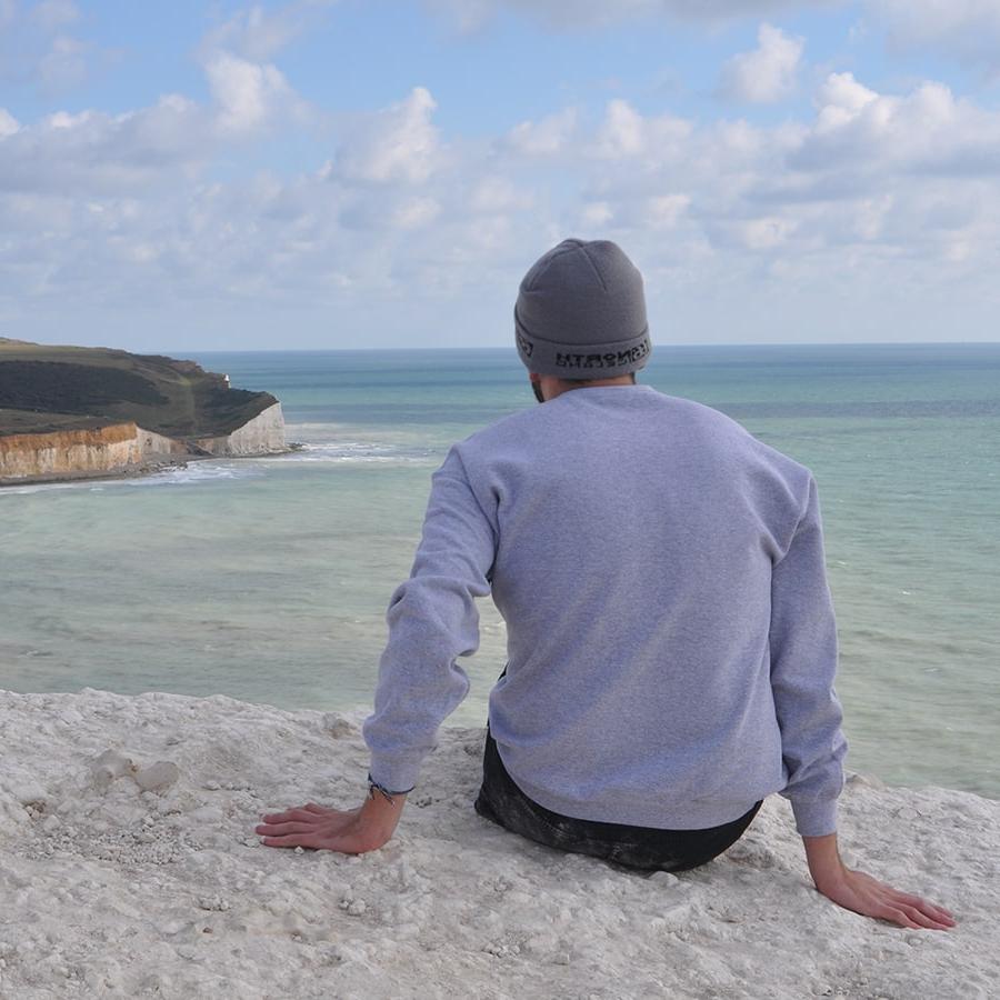 Student sits on beach in long sleeve shirt staring out to sea.