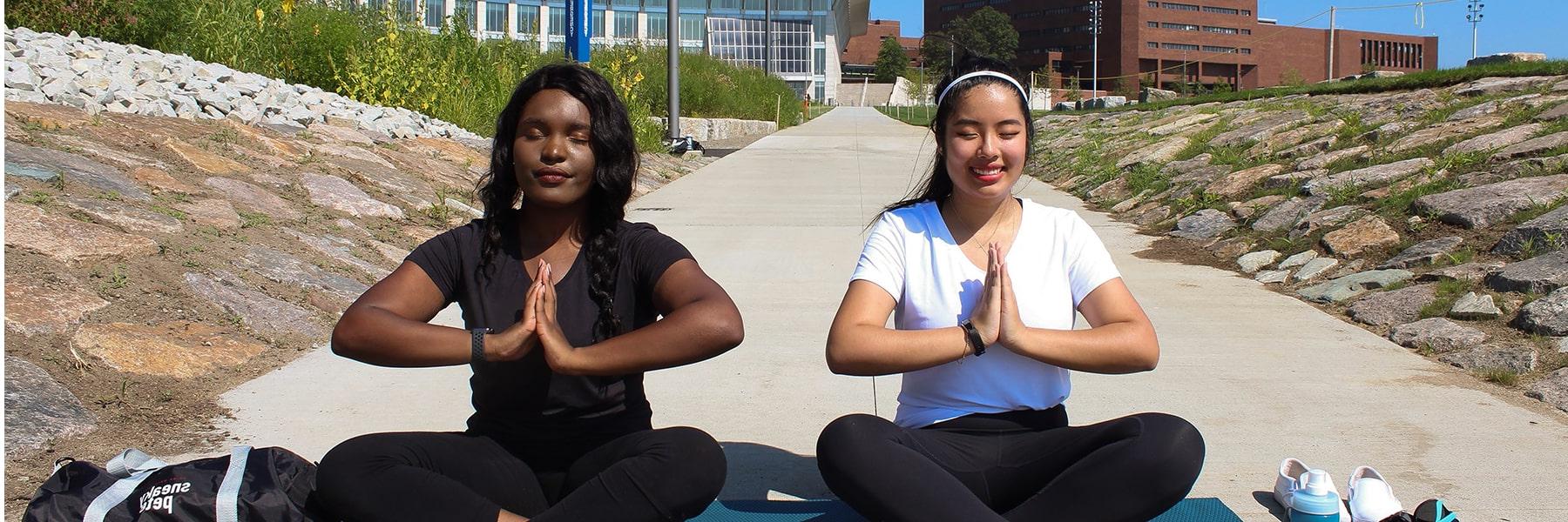 校园中心前，两名学生盘腿而坐，双手并拢，摆出瑜伽姿势.